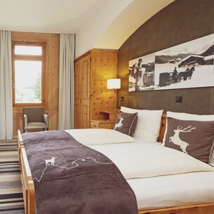 Double room with deer-style bedding, window with chair and wooden furnishings | © Davos Klosters Mountains 